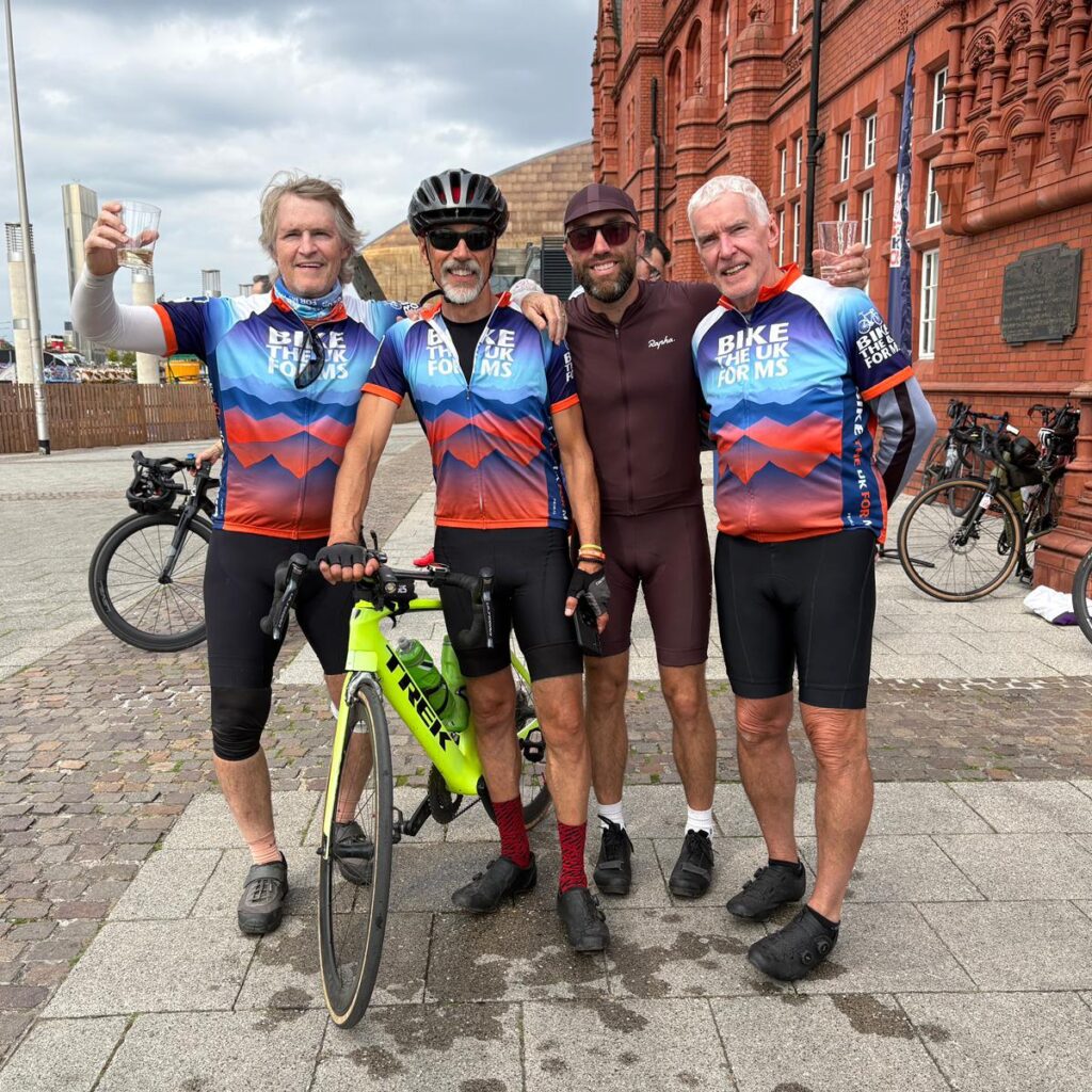 4 men and a bike smiling for the camera, arms around each other and cheering a class of champagne having completed Lon Las Cymru 